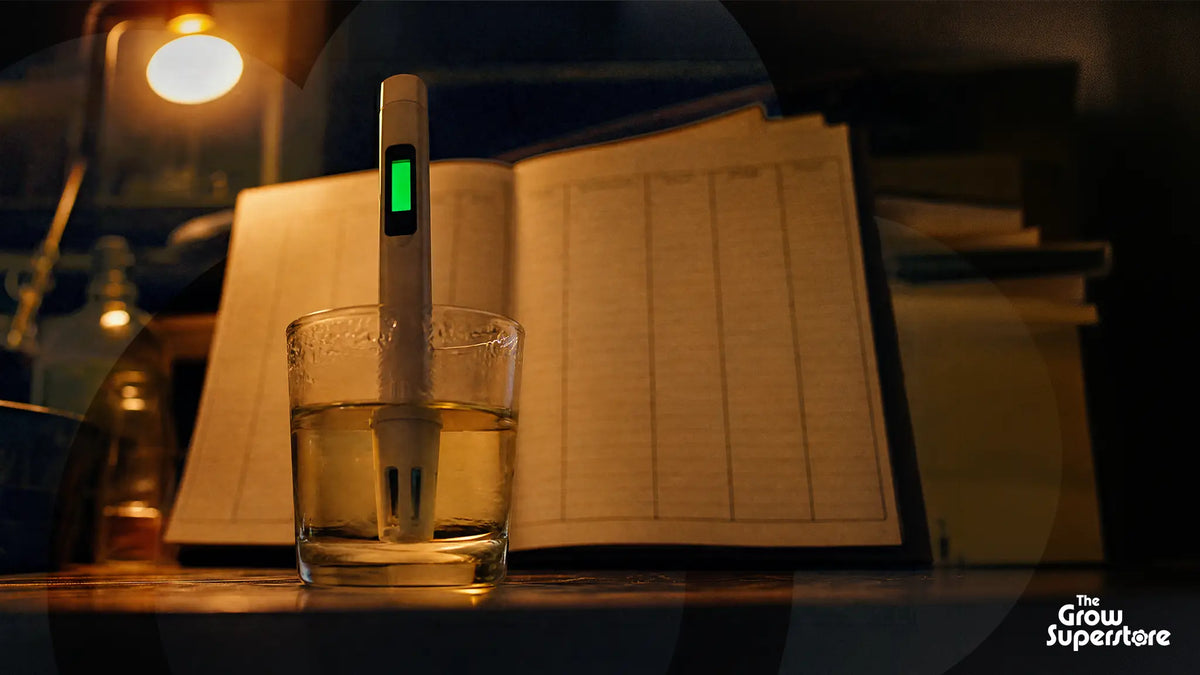 White EC meter with green display standing in a glass of nutrient solution beside an open feeding log book in warm lamplight