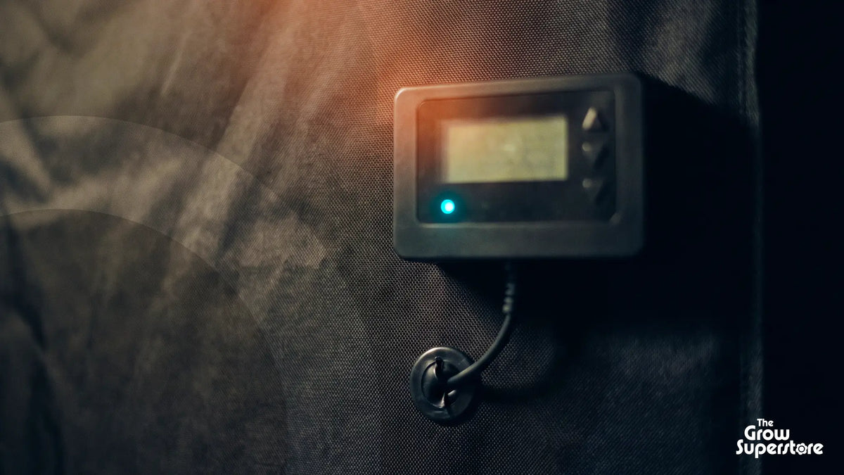 Grow room controller with LCD screen, arrow buttons and glowing blue light on dark grow tent fabric, cable through a port